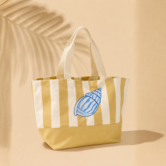 A canvas tote bag with vertical cream and mustard yellow stripes, a solid mustard yellow bottom, and two cream fabric handles. A blue seashell graphic is printed on the front.