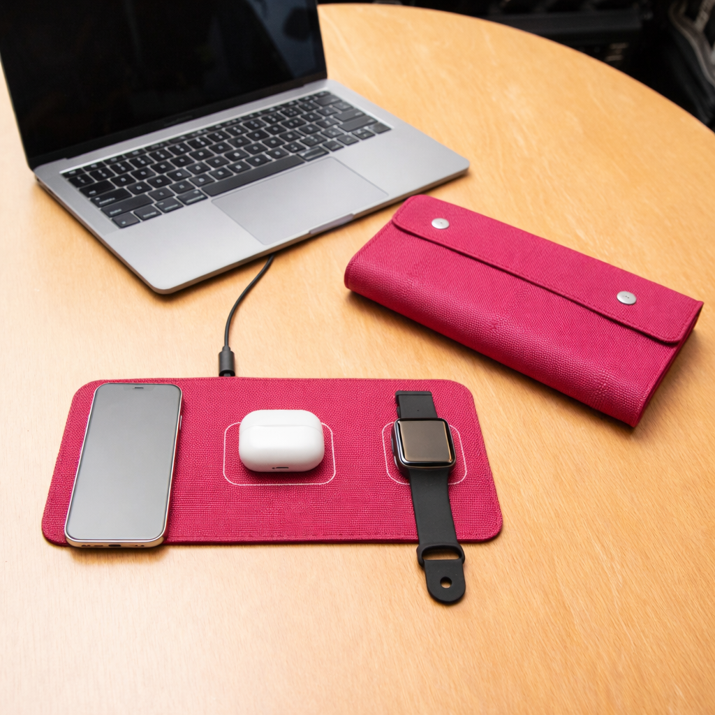 Pink charging pad with smartphone, white mouse, and smartwatch on a wooden table with a laptop.