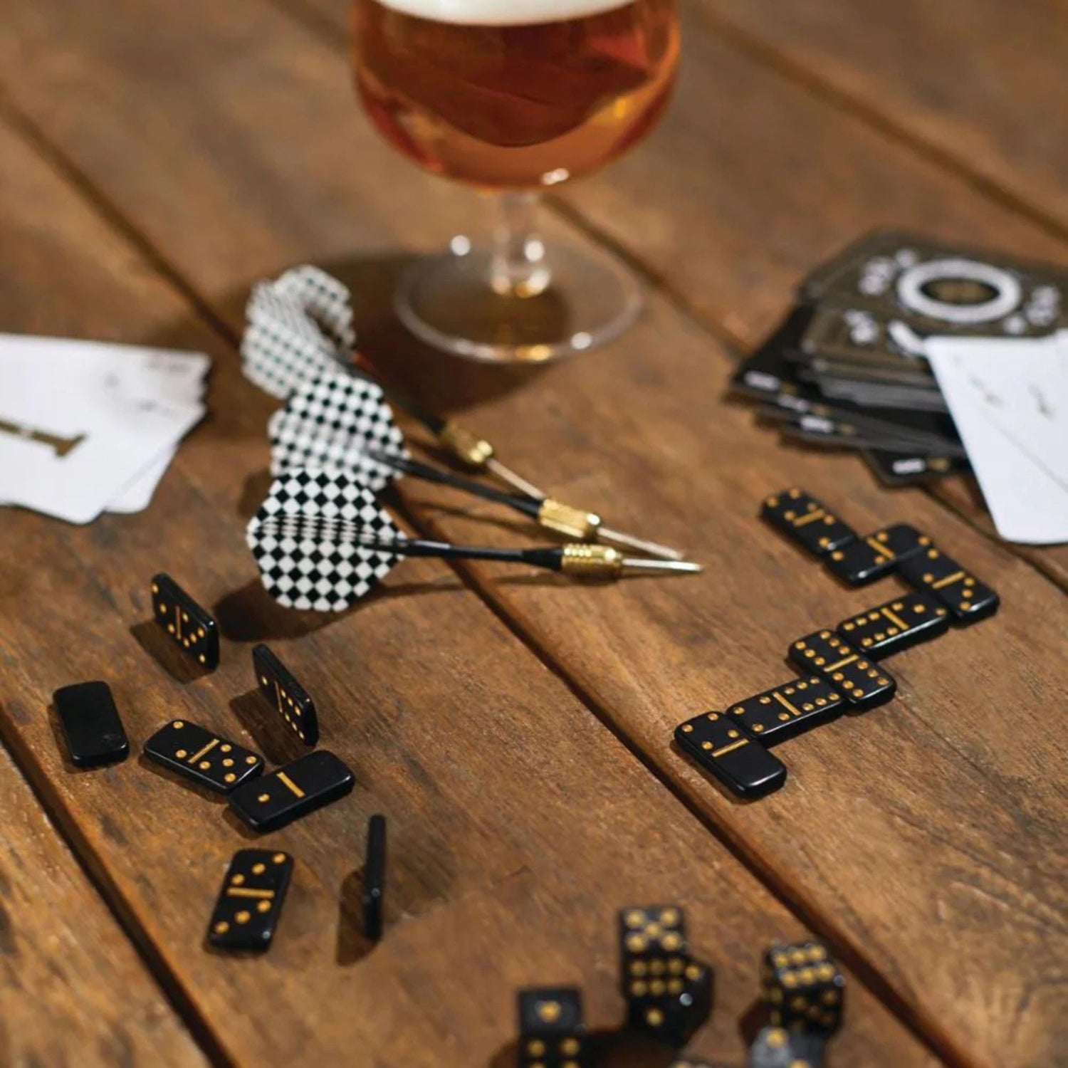 Black and gold dominoes with black-and-white checkered darts arranged on a rustic wooden table, alongside playing cards and a glass of amber beer.