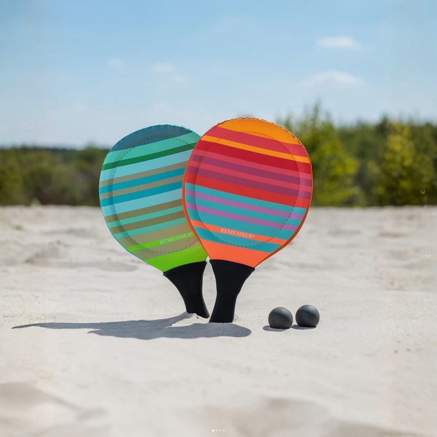 Two colorful striped beach paddles with two balls on a sandy surface.