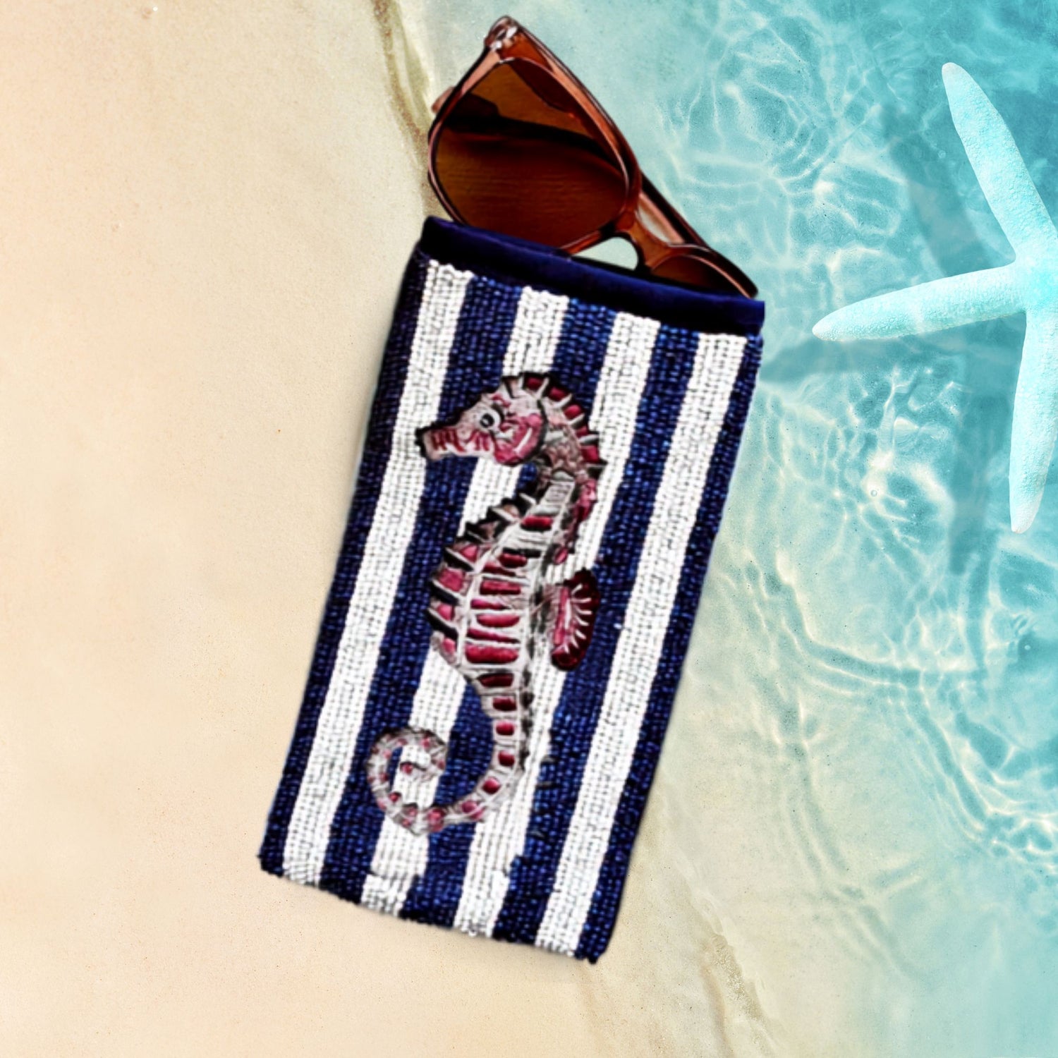 Striped sun glass case with seahorse design containing sunglasses, on a beach setting.
