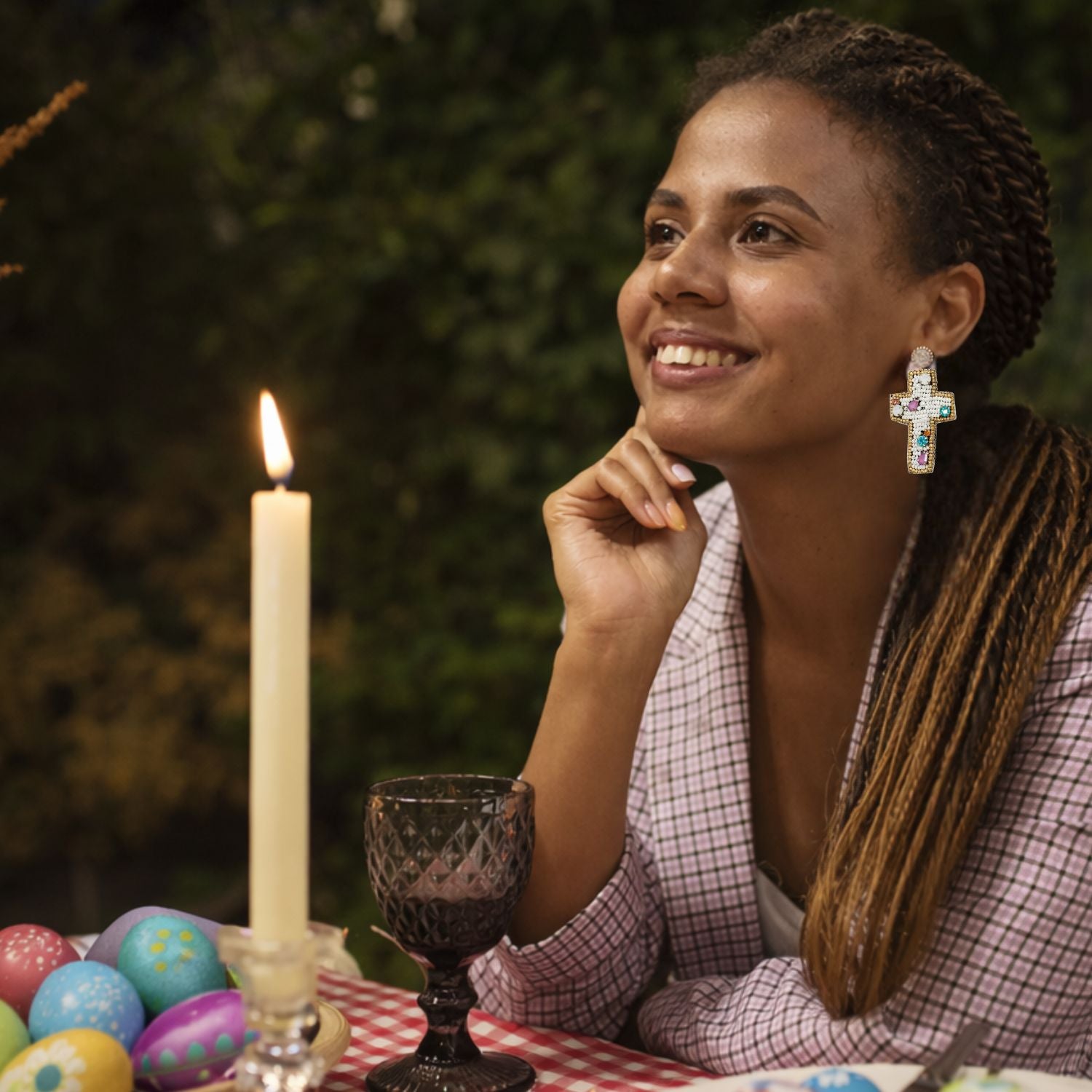 Woman with beaded Easter earrings sitting at a table with Easter eggs and a lit candle, smiling outdoors.