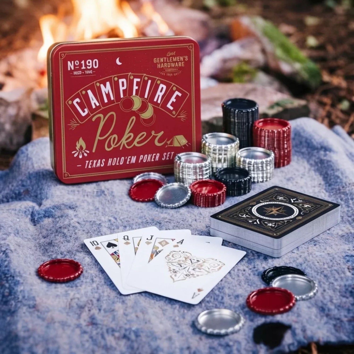 Campfire Poker Texas Hold'em poker set displayed outdoors on a blue blanket with a campfire in the background. The set includes a red tin box, a deck of cards, several silver and red bottle caps, and stacks of poker chips.