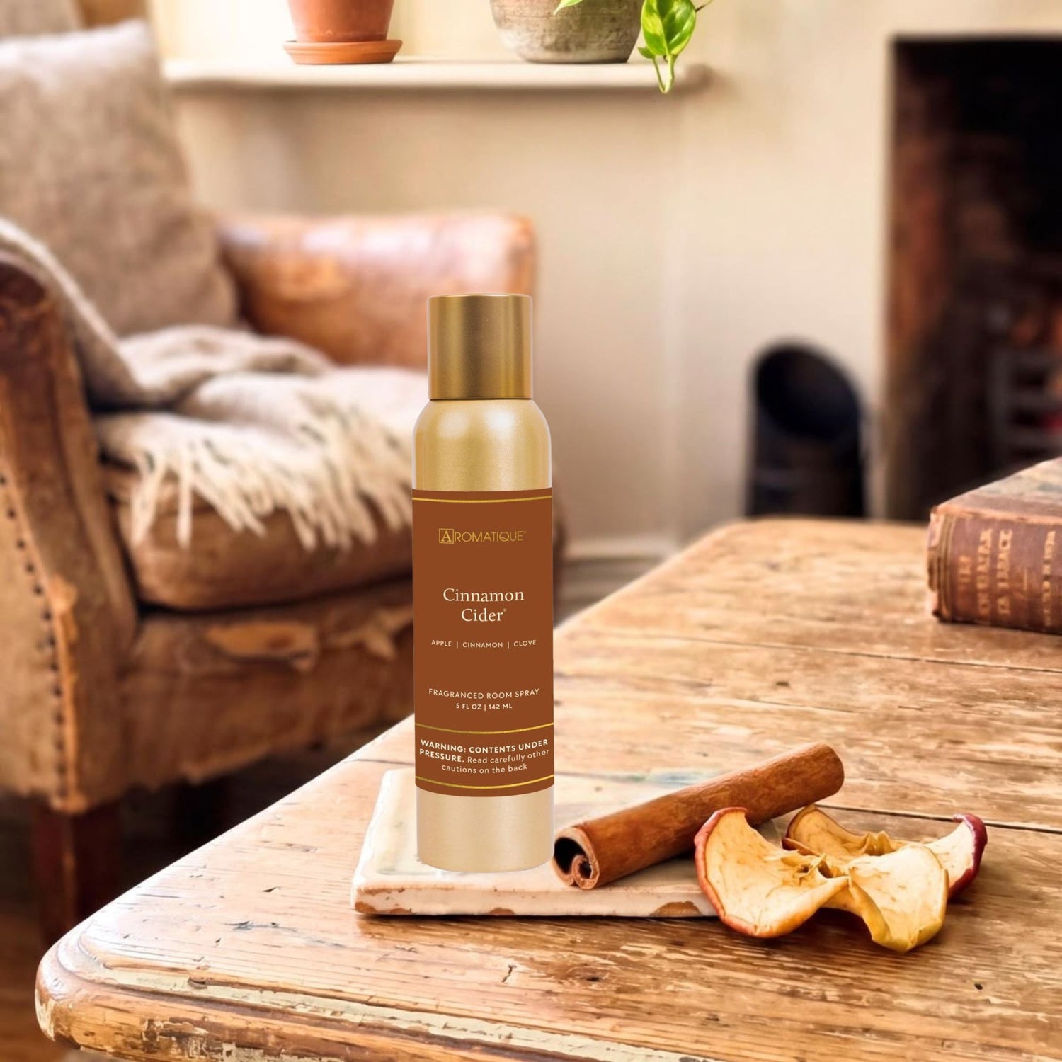 A bottle of 'Cinnamon Cider' room spray on a wooden table with cinnamon sticks and dried apples in a cozy living room setting.