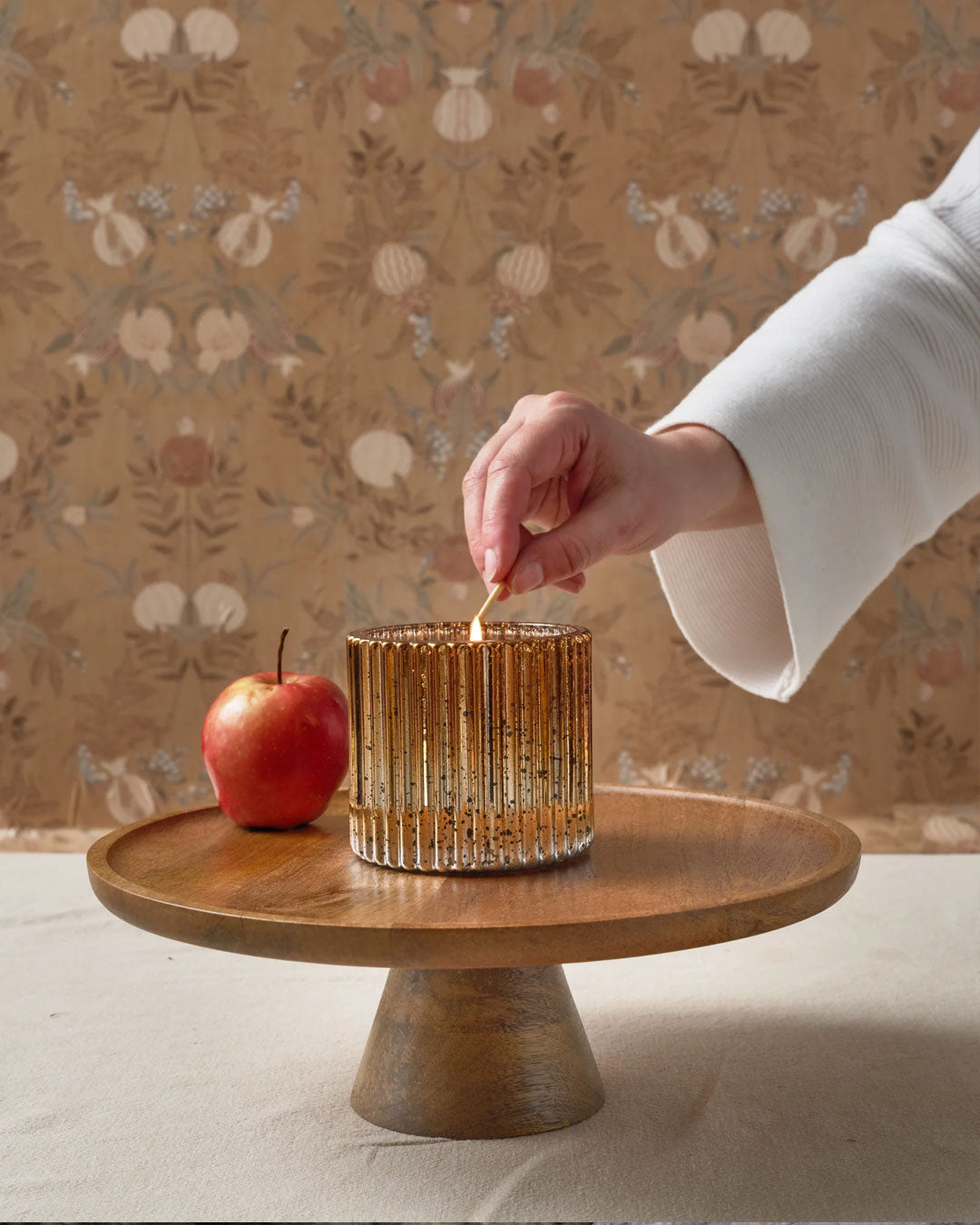 Hand lighting a cinnamon spice scented candle on a wooden stand with an apple, against a decorative wallpaper background.