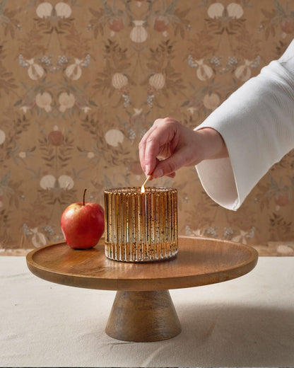 Hand lighting a cinnamon spice scented candle on a wooden stand with an apple, against a decorative wallpaper background.