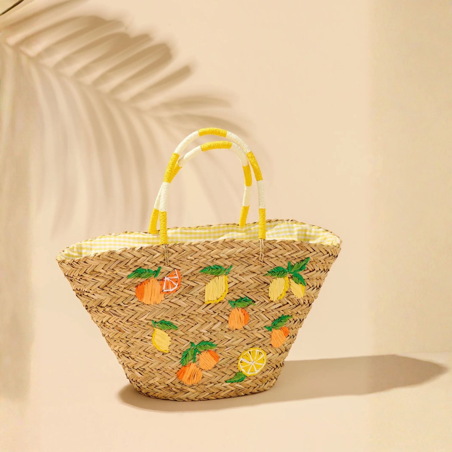 A woven straw tote bag with yellow and white striped handles, decorated with embroidered pineapples and lemons on the front, set against a warm beige background with soft palm leaf shadows.