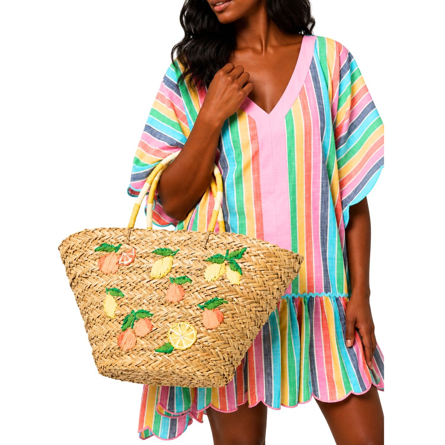 A woman wearing a pastel rainbow striped kaftan dress and holding a large woven straw tote bag decorated with embroidered citrus fruits, set against a plain white background.