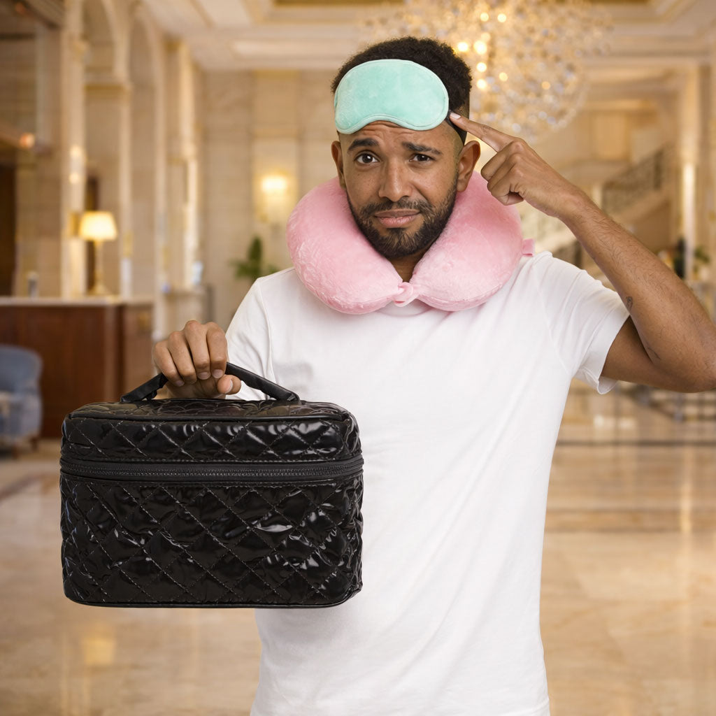 Man holding a black travel case with a pink neck pillow and green sleep mask in a hotel lobby.