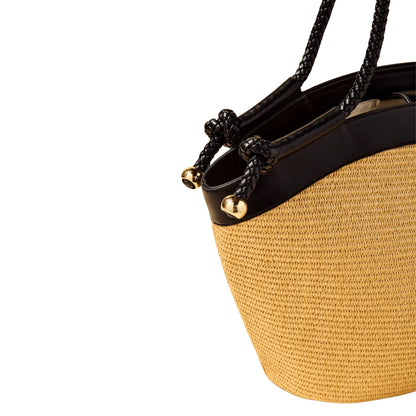 Close-up of a woven straw bucket bag with black leather trim, black braided shoulder straps, and small gold bell accents on a white background.