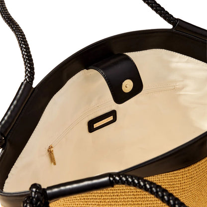 Close-up of the interior of a woven straw tote bag with black leather trim, braided black handles, a cream-colored lining, and a small gold-tone metal brand plaque.