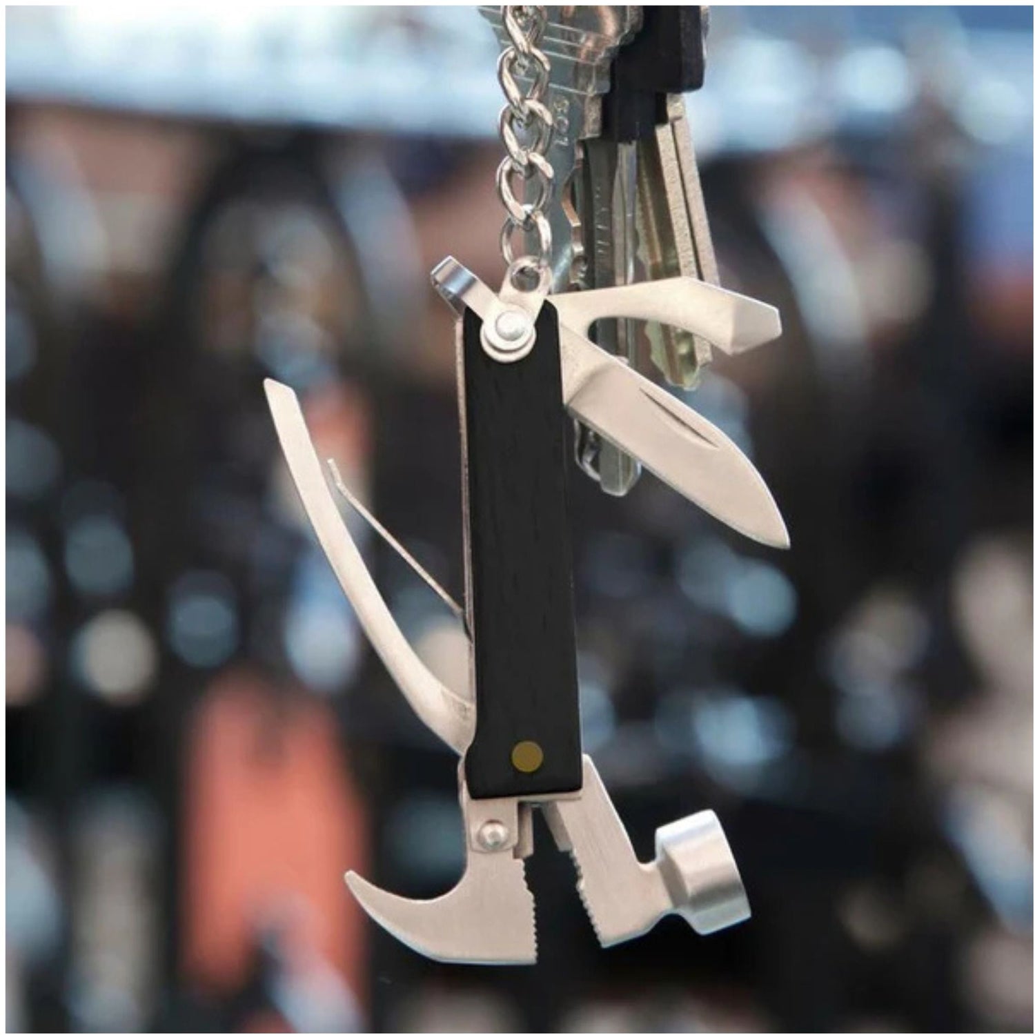 A multi-tool keychain with a black handle and silver metal tools including a knife, axe, and hook, attached to a silver keyring with keys, shown against a blurred dark background.