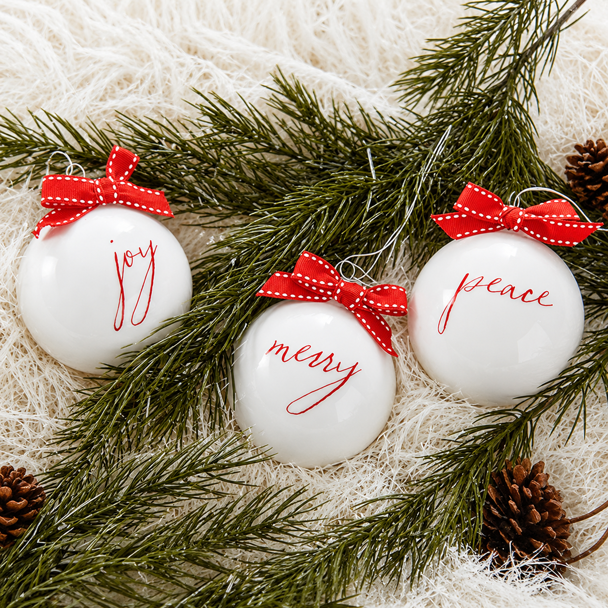 Set of three white glass Christmas ornaments with Joy, Merry and Peace lettering and red ribbon bows nestled among pine branches.