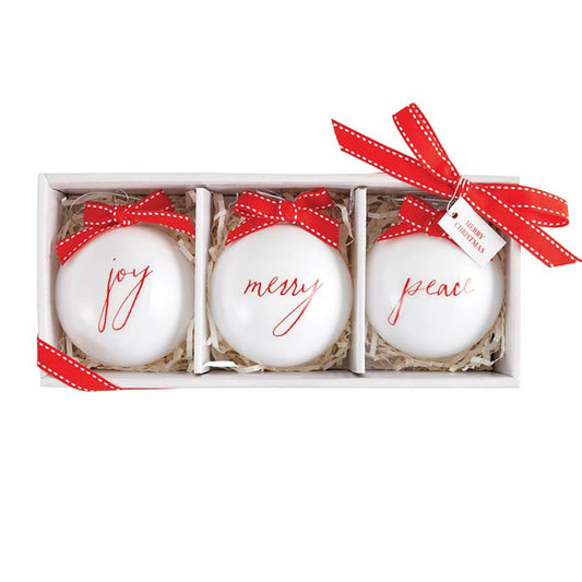 Set of three white glass Christmas ornaments with Joy, Merry and Peace lettering and red ribbon bows nestled among pine branches.