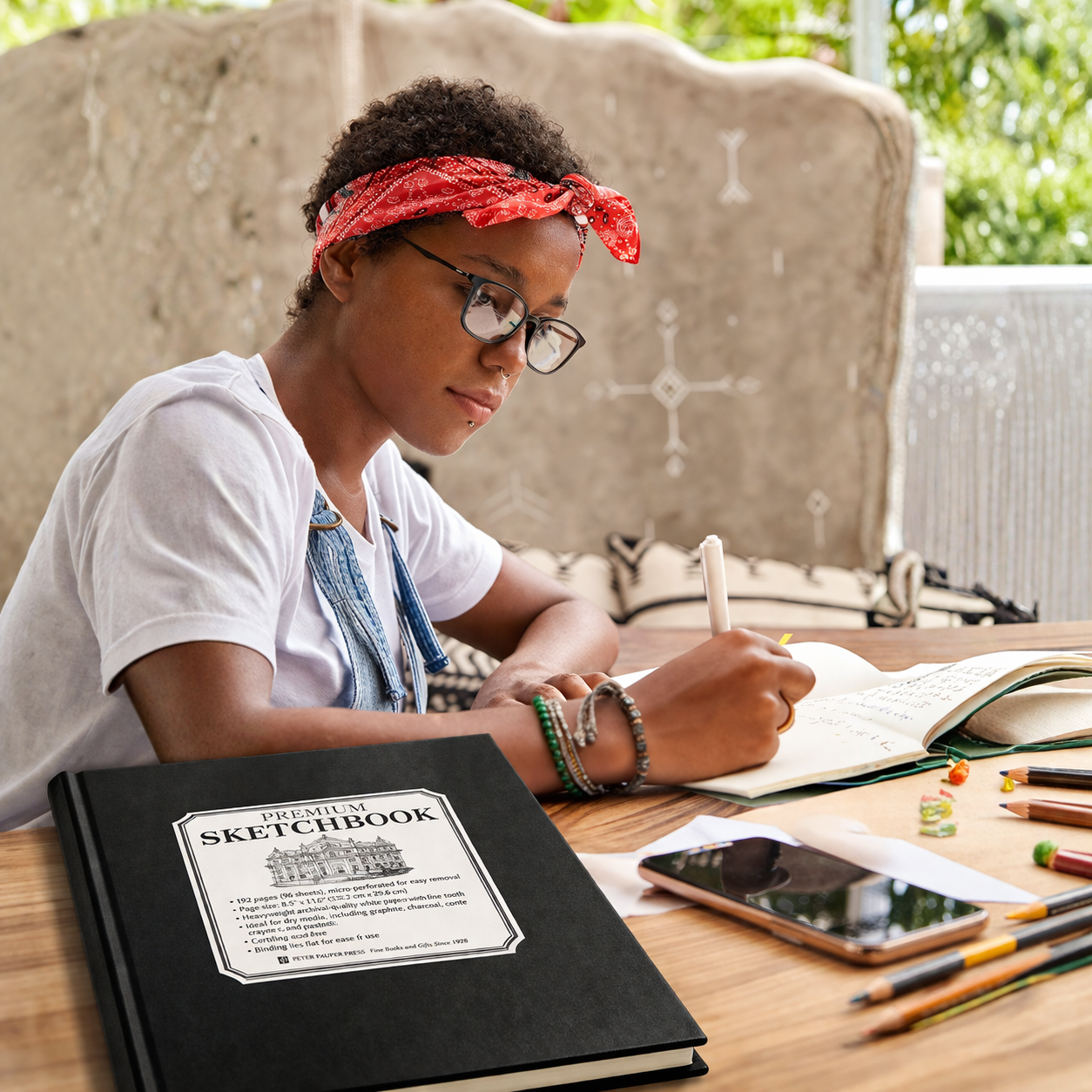 Young Student sitting at a table with a sketchbook and art supplies, wearing a red bandana and glasses.