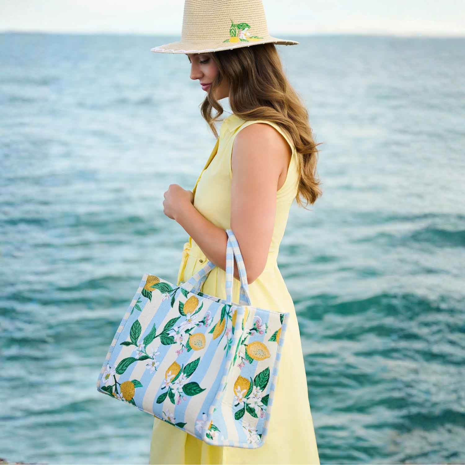 Woman in a yellow dress and hat holding a tote bag with lemon design by the ocean.