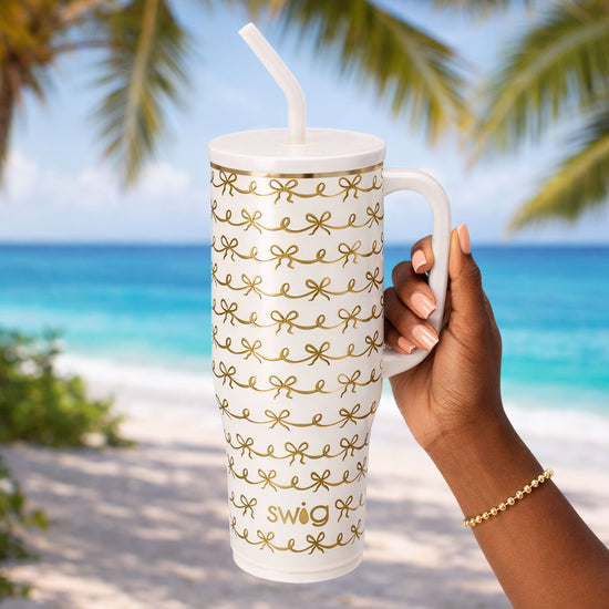 Hand holding a white mug with gold patterns and 'Swig' branding on a beach with palm trees.