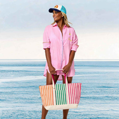 Woman in pink cover-up and colorful cap holding a striped beach bag on a beach.