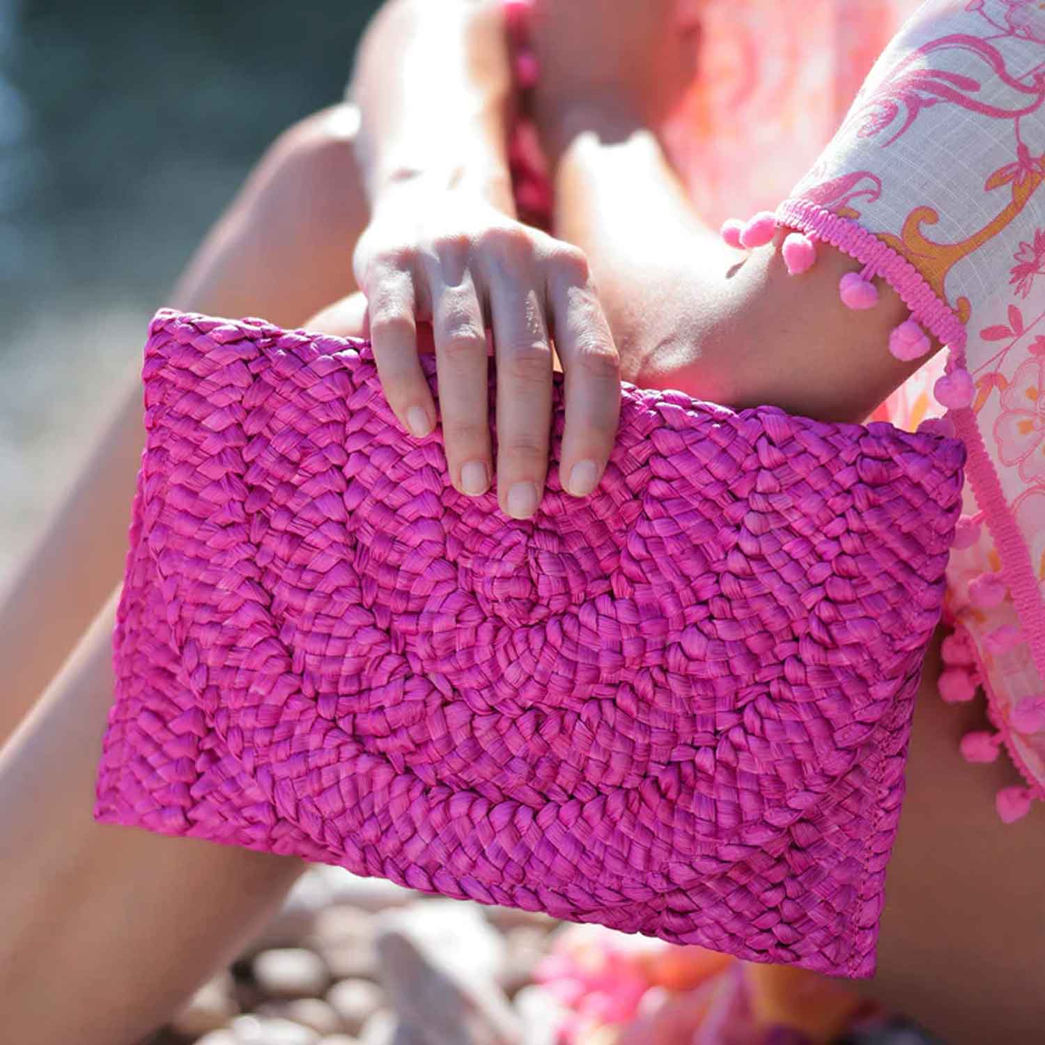 Woman sitting on a beach holding a pink woven clutch with blurred background