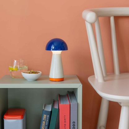 Colorful mushroom-shaped lamp on a shelf with books and a chair against a peach wall.