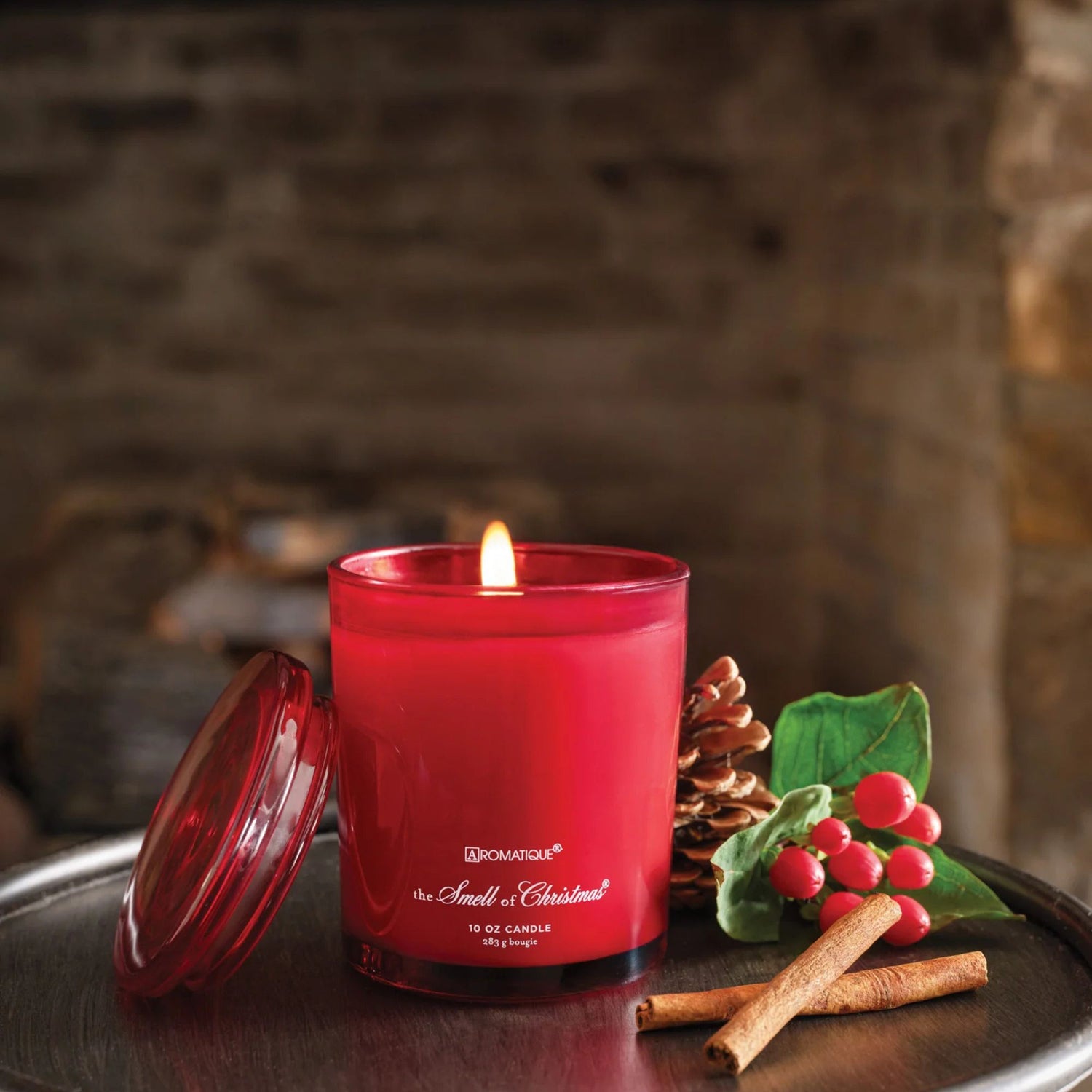 Red candle with a lit wick on a dark surface, surrounded by cinnamon sticks and holly leaves.