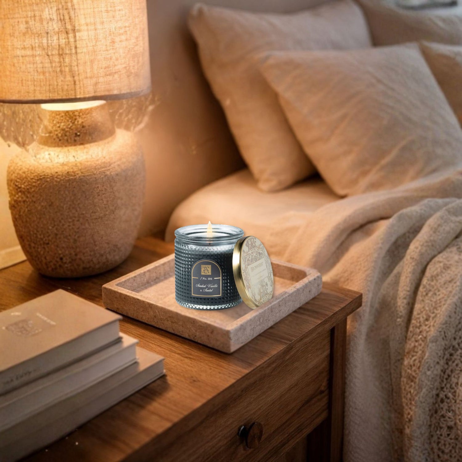 Vanille Santal Candle on a nightstand next to a lamp and books, with pillows in the background