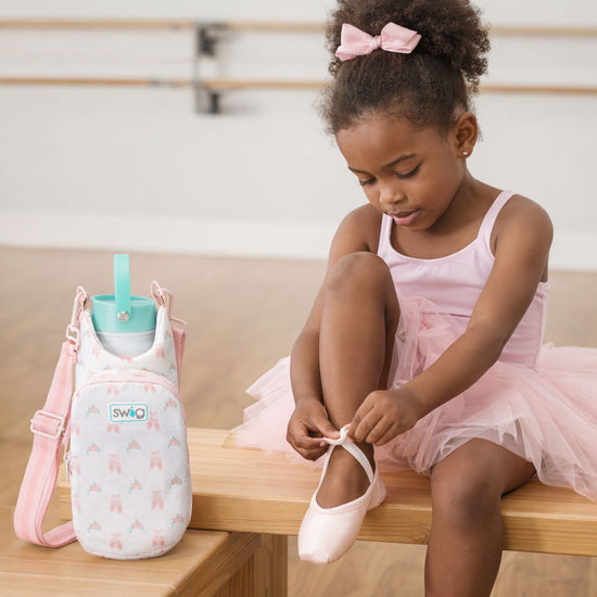 Young girl in a pink tutu sitting on a bench with a cute bottle and bag next to her.